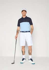 Tengerészkék és világoskék galléros póló fehér rövidnadrággal párosítva. Kiegészítők: sötét öv és fehér cipők, kezében golfütő.