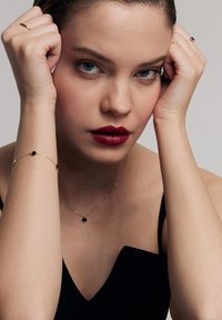 Czarny naszyjnik z okrągłym wisiorkiem i pasującą bransoletką, obie na złotych łańcuszkach. Modelka ma wysoko związane włosy w kok, nosi czarną bluzkę i ciemną pomadkę.