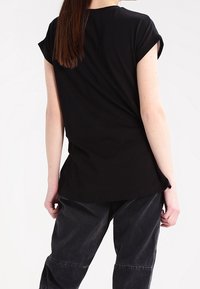 Persona con cabello largo y marrón, vistiendo una camiseta negra de manga corta y pantalones negros holgados, de pie de espaldas frente a un fondo blanco.