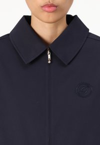 Jachetă navy cu un guler mare, închidere cu fermoar și detalii cu logo embosat. Fabricată dintr-un material neted cu o culoare solidă și accente minime.