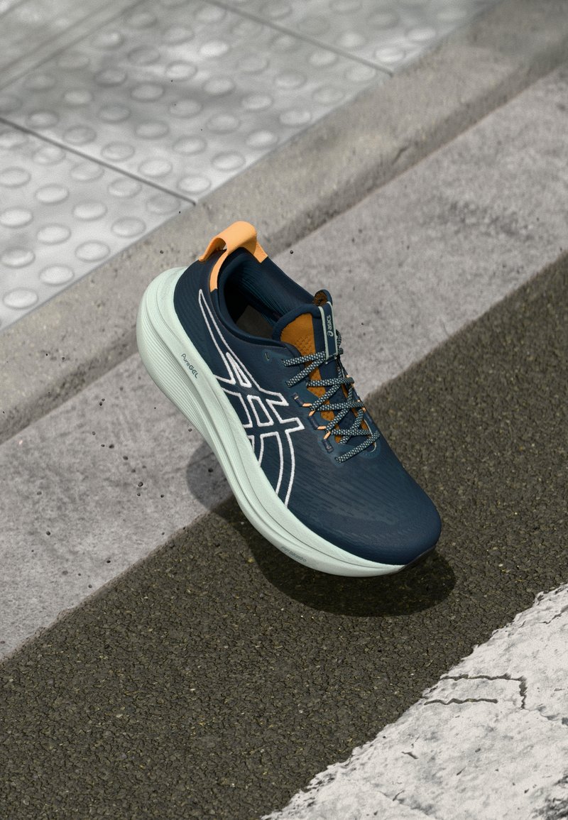 Chaussure de sport avec une tige en maille bleu marine, une semelle intermédiaire vert clair, un onglet de talon orange et une marque blanche. Les lacets et la texture offrent un design moderne.