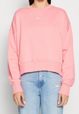 Kobieta ubrana w jasnoróżową krótką bluzę Nike z długimi rękawami i jasnoniebieskie podarte jeansy na jednolitym tle.