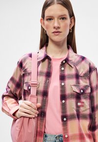 Camisa xadrez de flanela em tons de rosa, bege e roxo, com botões e um bolso no peito. Combinada com uma t-shirt rosa e uma mala de alça rosa.