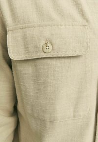Tessuto beige leggero con finitura testurizzata, caratterizzato da una tasca con chiusura a bottone rotondo nella zona del petto.