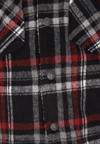 Camisa de franela a cuadros en negro, gris y rojo, con dos bolsillos en el pecho y botones metálicos con detalle de logo. Tejido suave y texturizado.