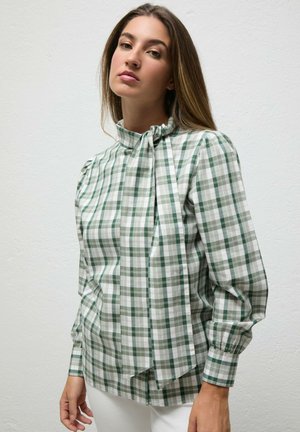 Joven mujer con el pelo largo, que lleva una blusa de cuadros verdes y blancos con un lazo alto en el cuello y pantalones blancos, de pie junto a una pared lisa.