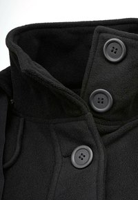 Col haut en polaire noire avec trois grands boutons noirs, texture douce et design légèrement surélevé. Comprend des poches latérales et une fermeture �éclair.