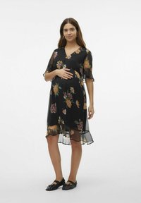 Zwarte doorschijnende zwangerschapsjurk met bloemmotief, V-halslijn, korte mouwen, asymmetrische zoom en vloeiende textuur. Gecombineerd met platte zwarte schoenen.