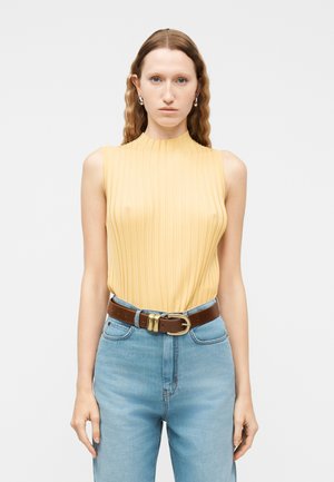 Kvinde med langt bølget hår iført en ærmeløs gul ribbet top, stoppet ned i højtaljede lyseblå jeans med brunt bælte og sølvøreringe.