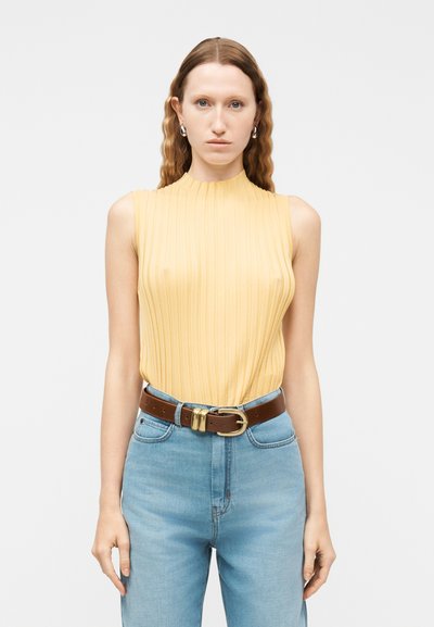 Femme aux longs cheveux ondulés portant un débardeur jaune côtelé rentré dans un jean bleu clair taille haute avec ceinture marron et boucles d'oreilles argentées.