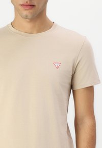 Homme portant un t-shirt beige à manches courtes avec un petit logo Guess rouge sur la poitrine gauche, présenté des épaules au haut du torse.