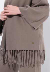 Poncho marrón con flecos, fabricado en material suave, que cuenta con mangas anchas y una pequeña etiqueta dorada cerca del dobladillo. Textura suave y corte relajado.