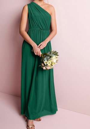 Vrouw draagt een groene, floor-length, een-schouderjurk en houdt een boeket bleekgele en roze bloemen vast tegen een lichtroze achtergrond.