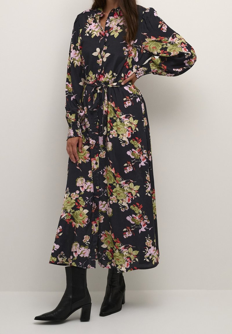 Femme portant une longue robe noire à fleurs avec une taille nouée et de longues manches bouffantes, associée à des bottines noires à talons.