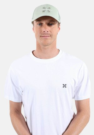 Homme portant une casquette vert clair avec un motif d'oiseau et un t-shirt blanc avec un petit logo noir sur la poitrine, sur un fond uni.