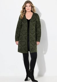 Olivgrüner Cardigan mit schwarzen Herzmustern, offene Front, lange Ärmel und weicher Textur, kombiniert mit schwarzen Leggings und Ankle Boots.