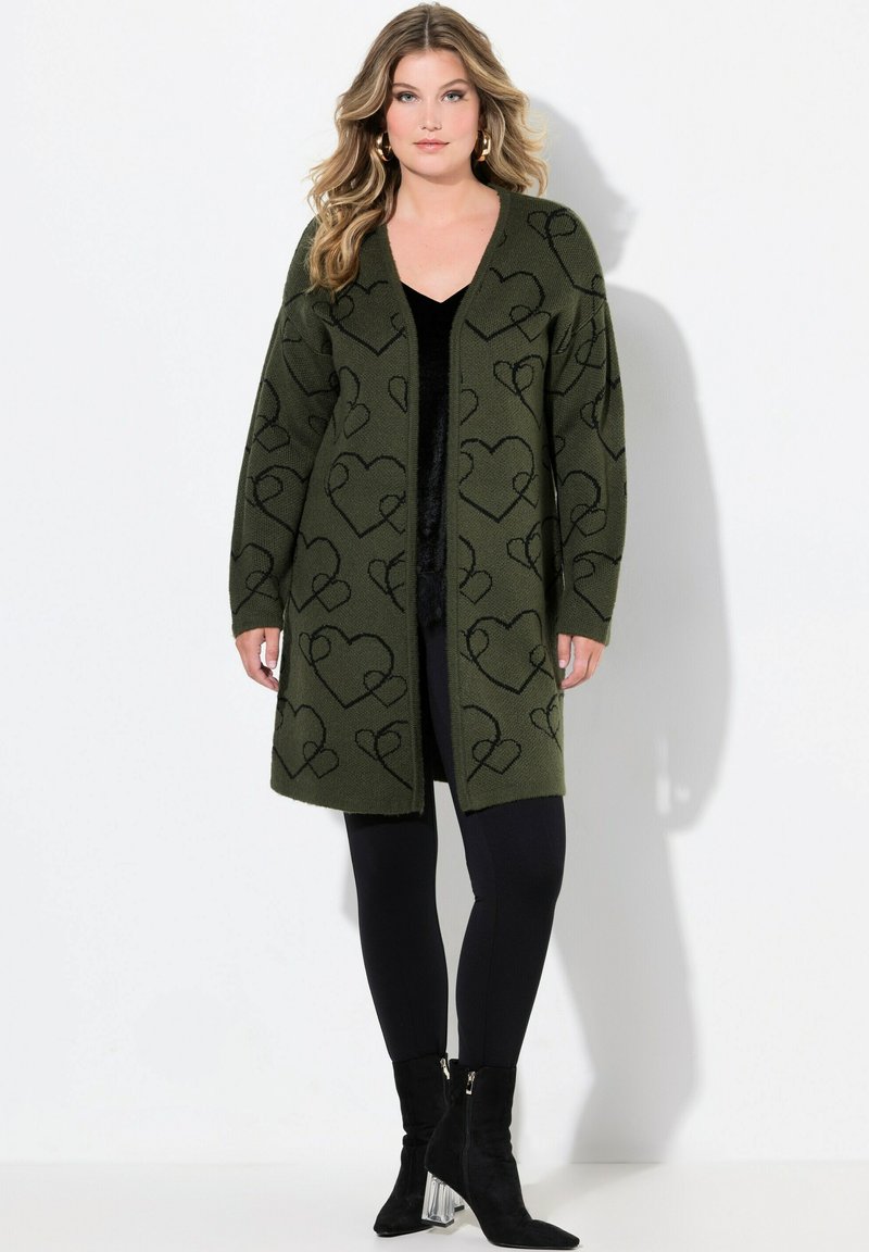 Olivgrüner Cardigan mit schwarzen Herzmustern, offene Front, lange Ärmel und weicher Textur, kombiniert mit schwarzen Leggings und Ankle Boots.