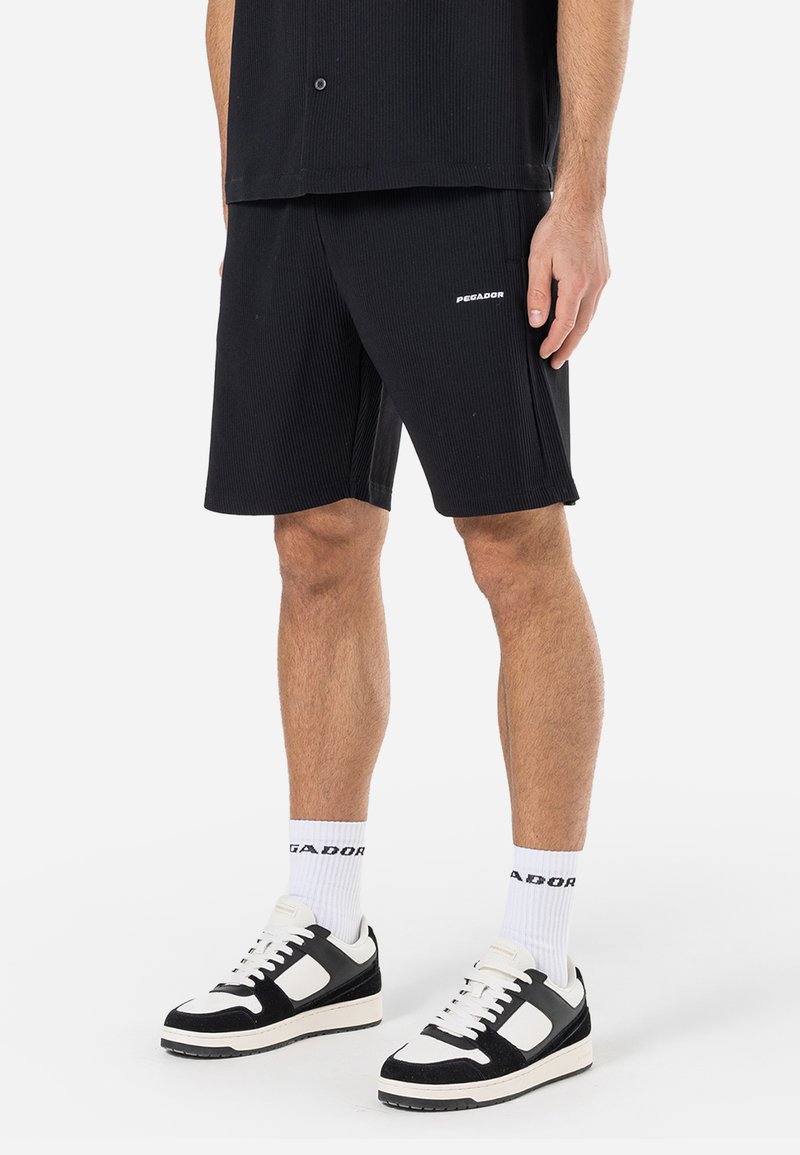 Shorts noirs à côtes en tissu texturé, avec des fentes sur les côtés et un petit logo blanc. Associés à des baskets noir et blanc et des chaussettes blanches.