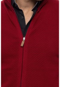 Chaqueta roja con cremallera que presenta un patrón de tejido texturizado y un tirador de cuero en la cremallera, llevada sobre una camisa negra con cuello redondeado.