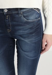 Jeans de denim azul oscuro con un ajuste slim, que cuentan con sutiles desgastes, bolsillos frontales y un acento de logo cerca de la cinturilla.