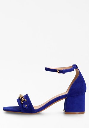 Sandales à talons en suede bleu avec une bride au niveau de la cheville, un design à bout carrée, des accents en métal doré et un talon bloc épais.