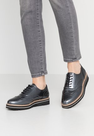 Osoba nosząca metaliczne czarne buty brogue z grubymi podeszwami i szare obcisłe dżinsy na gładkiej białej podłodze.