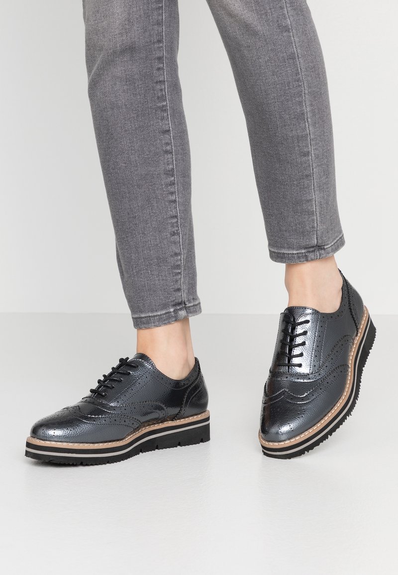 Personne portant des chaussures brogue noires métalliques à semelles épaisses et un jean skinny gris sur un sol blanc uni.