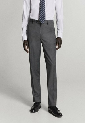Hombre con pantalones de vestir grises, camisa blanca, corbata navy con rayas y zapatos negros de vestir, de pie frente a un fondo liso.