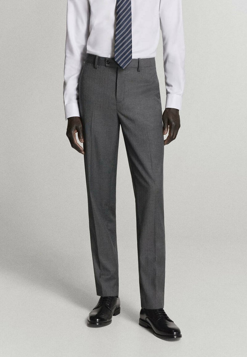 Homme portant un pantalon de costume gris, une chemise blanche, une cravate rayée bleu marine et des chaussures habillées noires, debout devant un fond uni.