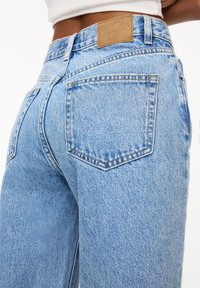 Jeans en denim bleu clair avec une taille haute, comportant deux poches arrière et une étiquette en cuir beige à la taille.