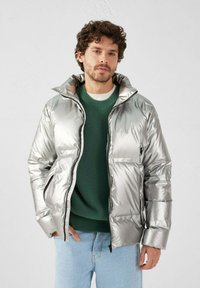 Giacca a soffietto argentata con una finitura lucida e riflettente; presenta un colletto alto, chiusura con zip e due tasche frontali, indossata sopra un maglione verde.