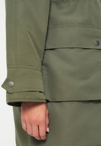 Olivengrøn jakke med en glat tekstur, der har en knappet manchet og et lagdelt design langs kanten. Bemærkelsesværdige detaljer i sølvhardware.