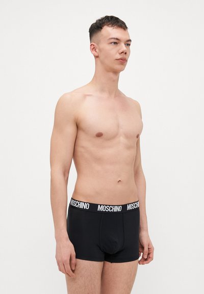 Black cotton boxer shorts with a white Moschino logo waistband, featuring a snug fit and smooth texture. Model stands in profile.