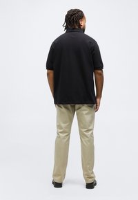 Άνδρας με πλεξούδες που φοράει μαύρο κοντομάνικο πόλο, μπεζ παντελόνι και μαύρα παπούτσια, ο οποίος στέκεται γυρισμένος με την πλάτη σε λευκό φόντο.