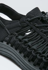 Black sandal featuring a synthetic upper with woven elastic cords. Textured sole with a thick design. Lightweight and open-toed.