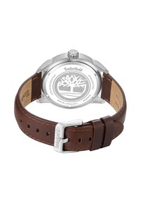 Timberland pulkstenis ar nerūsējošā tērauda korpusu, brūnu ādas siksniņu un metāla sprādzi. Aizmugurē ir koka logotips.