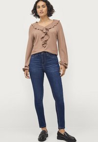Bruine geribbelde langemouwen top met ruches bij de hals, gecombineerd met donkerblauwe skinny jeans. Model draagt zwarte instappers.