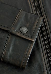 Poignet de veste en cuir noir avec des coutures visibles, un bouton rond noir portant l'inscription "Signature Durable Goods" et une fermeture éclair verticale à proximité.