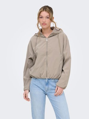 Jeune femme portant une veste à capuche beige clair zippée et un jean bleu clair, posant avec une main dans la poche sur un fond blanc.