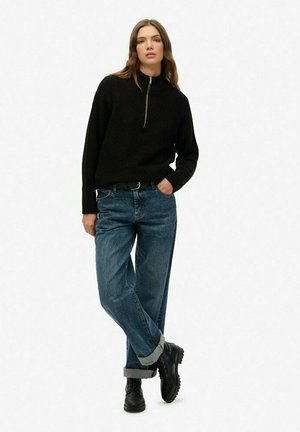 Zwarte, ritstrui met een hoge kraag; losvallende blauwe jeans met opgerolde boorden; zwarte veterschoenen; soepele textuur en ontspannen ontwerp.