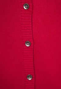 Tissu tricoté rouge avec une patte de boutonnage verticale côtelée et quatre boutons gris visibles cousus avec du fil rouge.