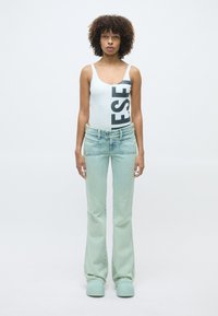 Body blanc avec des lettres noires, jean évasé en délavé clair et chaussures vert menthe. Le mannequin se tient face à un fond uni.