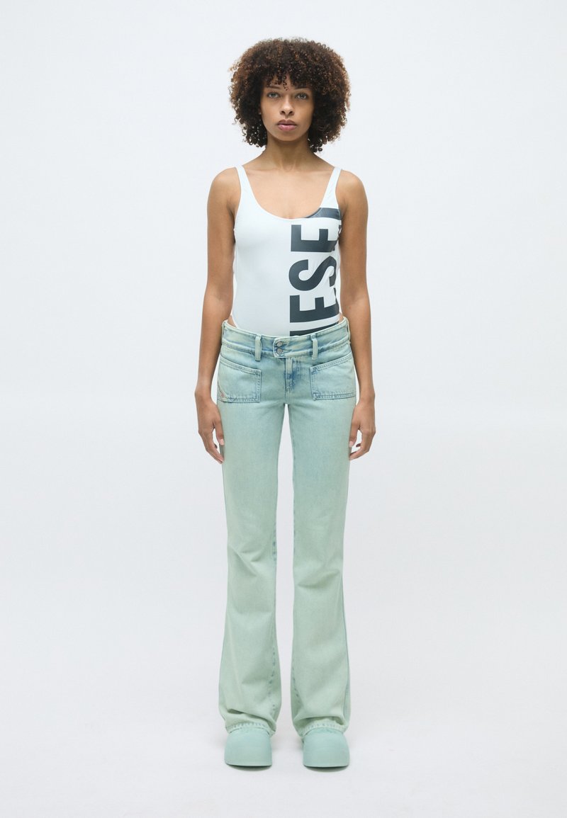 Body blanc avec des lettres noires, jean évasé en délavé clair et chaussures vert menthe. Le mannequin se tient face à un fond uni.