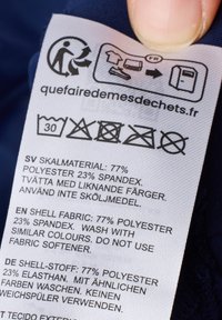 Etykieta z instrukcjami prania, skład tkaniny: 77% poliester, 23% elastan. Zawiera symbol recyklingu oraz wiele języków. Biały tekst na tkaninie.