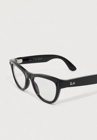 Gafas de sol Ray-Ban negras con montura de plástico gruesa, lentes transparentes y detalle del logo en las varillas. Vista en ángulo que muestra la forma y el diseño.