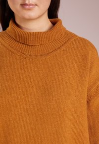 Jeune personne portant un pull à col roulé épais, côtelé et jaune moutarde, sur un fond uni pâle.