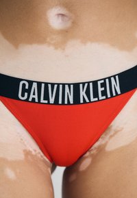 Rde bikinijevo dno z črnim elastičnim pasom, na katerem je v belem napisu "CALVIN KLEIN". Gladka tekstura, prilegajoč dizajn in minimalna pokritost.