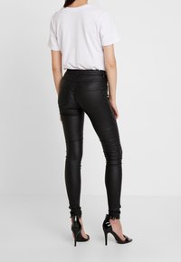 Femme portant un pantalon skinny en faux cuir noir, un t-shirt blanc à manches courtes et des chaussures à talons ouverts noires, se tenant devant un fond blanc.