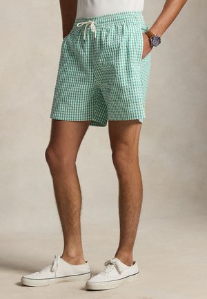 Uomo che indossa pantaloncini a quadretti verdi e bianchi con fascia elastica in vita, scarpe da ginnastica bianche e un orologio al polso sinistro.