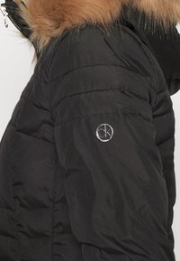 Svart puffjacka med en huva med pälsdetalj, quiltad textur och en framträdande silverfärgad CK-logotyp på ärmen. Mjuk material.